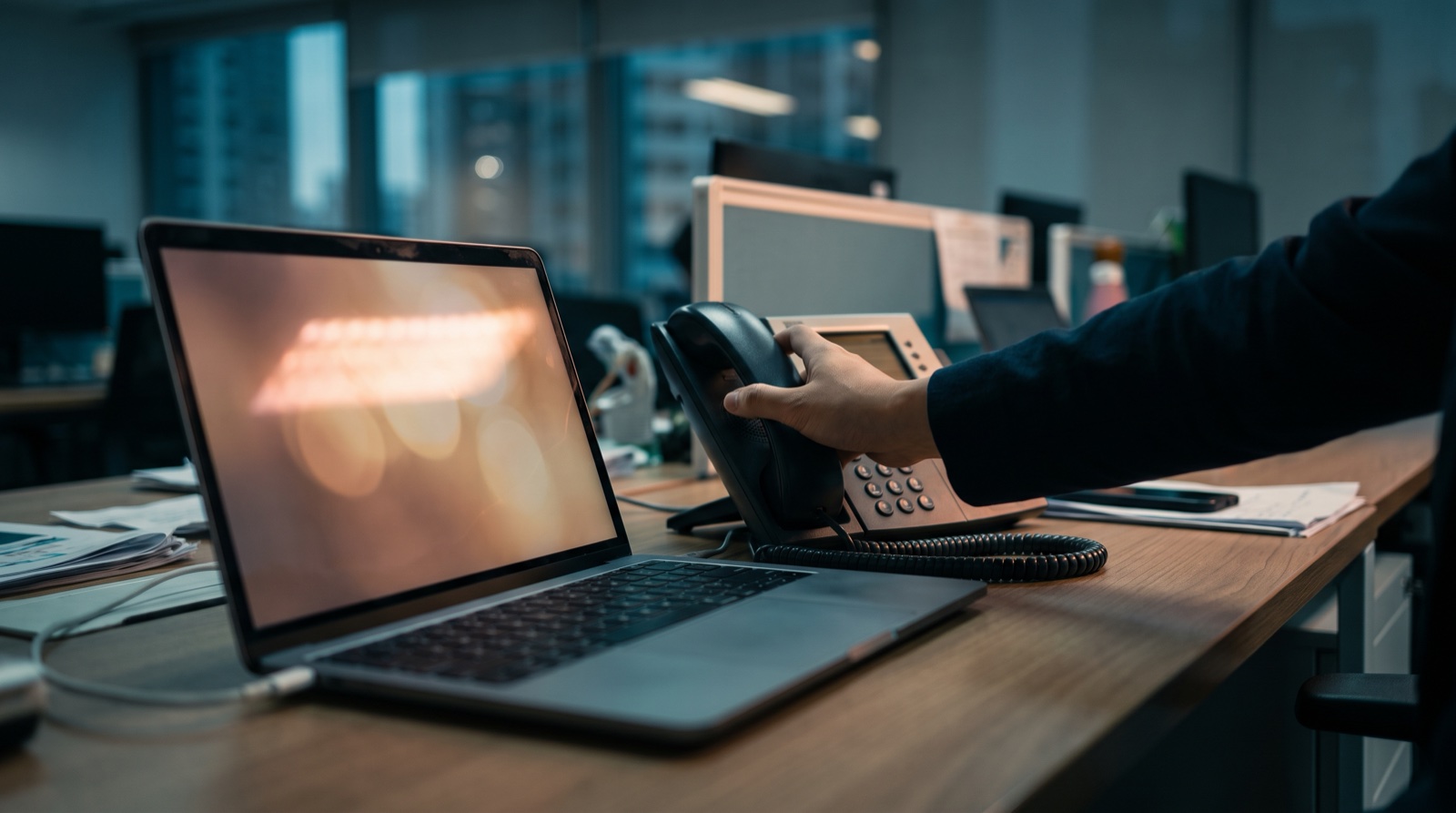 Photograph of a hand picking up a desk phone beside an open laptop with an unreadable, glowing screen, no face visible